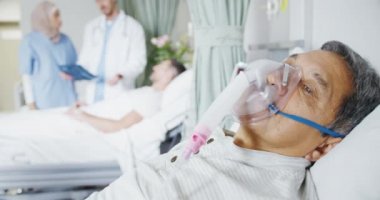 Hastane yatağında oksijen maskesi takmış son sınıf bir erkek hastayı yakından çek. Arka planda iki doktor başka bir hastayla konuşuyor. Önplana odaklan. 
