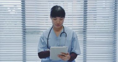 Genç Asyalı kadın doktorun tablet kullanırken ve muayenehanedeki kamerayla konuşurken belini kaldır. 