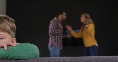 Beyaz çocuğun kanepeye yaslanması, anne ve babaların evde arka planda tartıştığı yakın çekim. Üzgün ve üzgün. 4k