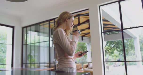 快適な家庭でコーヒーを飲み キッチンで携帯電話を使用して白人女性の側面図 彼女はテキスト4Kをテキストメッセージです — ストック動画