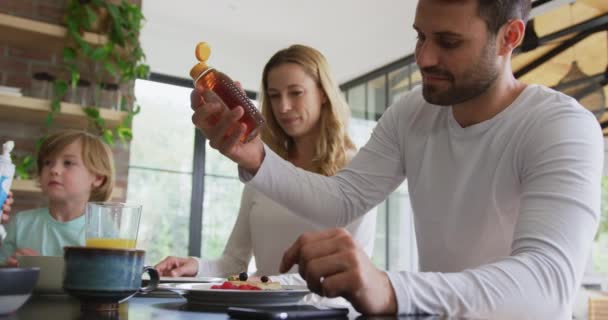 快適な家の台所でダイニングテーブルで食べ物を持っている白人の家族の低角度の眺め 食品4Kに蜂蜜を入れる父 — ストック動画