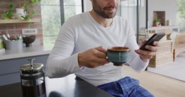 Beyaz adamın ön görünümünde kahve içmek ve rahat bir yemek masasında cep telefonu kullanarak. 4k mesaj atlıyor.