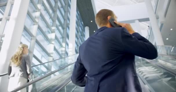 オフィスのエスカレーターで2階に移動し 携帯電話で話すビジネスマンの背面ビュー — ストック動画