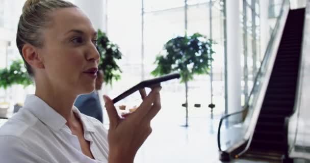 オフィスのロビーでエスカレーターの近くに立っている間 携帯電話で話している白人のビジネスマンの女性の側面図 彼女は離れて見ています4K — ストック動画