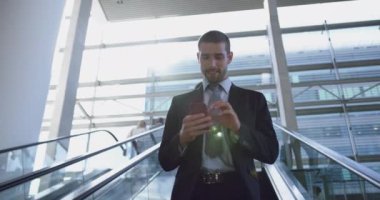 Kafkasyalı iş adamının modern bir ofiste cep telefonunu yürüyen merdivende kullanmasının düşük açılı görüntüsü. Gülümsüyor.