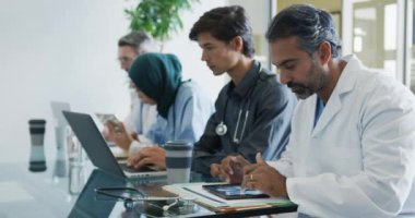 Bir masada birlikte çalışan çeşitli doktorların el kamerasıyla çekilmiş bir fotoğrafı. Orta yaşlı melez erkek doktor ön planda dönüp kameraya gülümsüyor