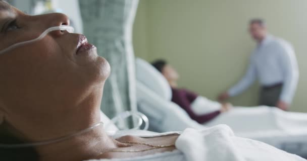 手持中年妇女混合泳特写 头戴鼻腹管躺在病床上 在后台接待来访者的病人 专注于前景 — 图库视频影像