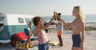 İki yetişkin kadın arkadaş, genç bir Kafkas ve genç bir Afro-Amerikan kadın Yan görünümü, bir tost yapma ve bir plajda birlikte duran bira şişeleri içme ve kucaklayan