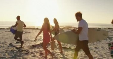 Gün batımında sahilde yürüyen, sörf tahtaları ve serin bir kutu taşıyan çok ırklı genç yetişkin arkadaşların yan görünümü. Arka -dan aydınlatmalı. Arkadaşlarla yaz eğlencesi, onlar plaj 4k bir yolculuğa vardır