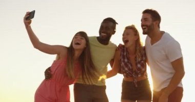 Gün batımında sahilde durup selfie çekerek çok ırklı bir genç yetişkin arkadaş grubunun portresini kapatın. Arka -dan aydınlatmalı. Arkadaşlarla yaz eğlencesi, onlar plaj 4k bir yolculuğa vardır