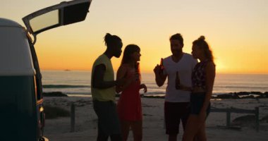 Siluet içinde genç yetişkin arkadaşlar bir yan görünümü onların kamp minibüsü tarafından konuşurken ve içme gün batımında bir plajda ayakta. Arkadaşlar 4k eğlenmek ile sahilde Yaz Sunset Partisi