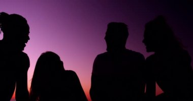 Gün batımında konuşan bir plajda oturan siluet genç yetişkin arkadaşlar çok ırklı bir grup yakından yan görünümü. Arkadaşlar 4k eğlenmek ile sahilde Yaz Sunset Partisi