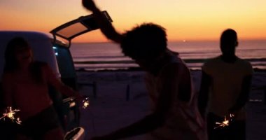 Gün batımında bir plajda siluet genç yetişkin arkadaş çok ırklı bir grup yakın, etrafında dans ve onların kamp minibüsü ile parlayan ışıltılar sallayarak eğlenin. Arkadaşlar 4k eğlenmek ile sahilde Yaz Sunset Partisi
