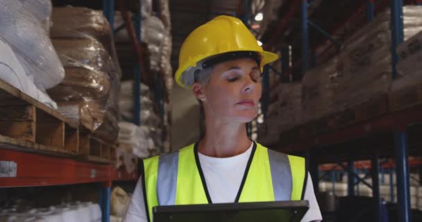 Vue rapprochée de face d'une travailleuse blanche d'âge moyen portant un casque jaune et utilisant une tablette pendant qu'elle patrouille dans les couloirs d'un entrepôt la nuit, levant les yeux vers la caméra et souriant 