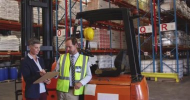 Orta yaşlı beyaz bir kadın ve orta yaşlı Kafkas erkek depo yöneticisi bir depo yükleme koylarında raflara işaret eden ve konuşurken bir forklift tarafından duran ön görünümü, o bir pano ve yazma tutuyor. 4k