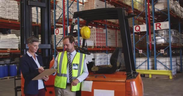 Vue de face d'une femme caucasienne d'âge moyen et d'un homme caucasien d'âge moyen gérant d'entrepôt debout près d'un chariot élévateur dans une baie de chargement d'entrepôt pointant vers des étagères et parlant, elle tient un presse-papiers et écrit. 4k 