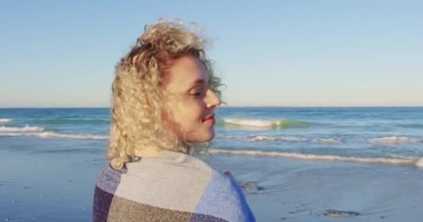 ショールを着て浜辺に立つ若い白人女性の横を見上げ 振り向いてカメラに向かって微笑み 海に向かって歩いて4K — ストック動画