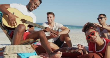 Yakın mutlu genç yetişkin erkek ve kadın arkadaş çok ırklı bir grup, birlikte genç bir Afro-Amerikan adam şarkı ve gitar çalarken dinleyerek bir plajda oturan. Genç arkadaşlar birlikte 4k plajda yaz eğlencesi