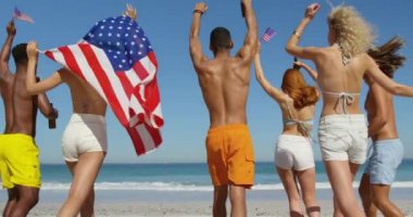 Sahilde dans eden, Amerikan bayraklarını sallayan ve kollarını havaya kaldıran çok ırklı genç yetişkin arkadaşların yakından görünümü. Genç arkadaşlar birlikte 4k plajda yaz eğlencesi