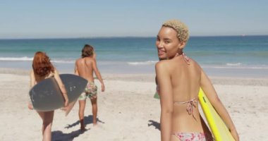 Bir sörf tahtası taşıyan arkadaşlarının arkasında bir plajda yürüyen genç bir karışık yarış kadın yakından arka görünümü, kameraya gülümseyerek etrafında dönerek ve çağırıyor. Genç arkadaşlar birlikte 4k plajda yaz eğlencesi