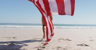 Bir Abd bayrağı 4k tutan denize doğru bir plajda çalışan genç bir Afrikalı-Amerikalı kadın düşük açılı arka görünümü