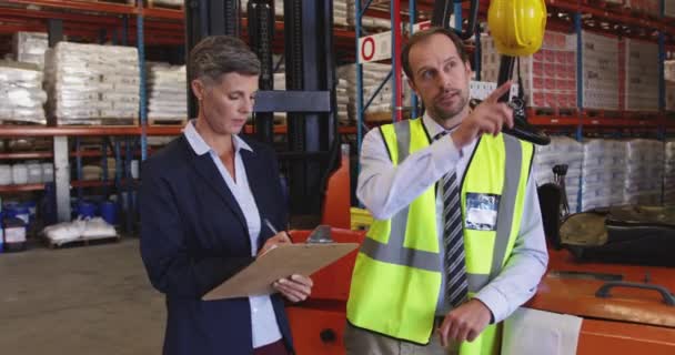Vue de face gros plan d'une femme caucasienne d'âge moyen et d'un homme caucasien d'âge moyen gérant d'entrepôt debout près d'un chariot élévateur dans une baie de chargement d'entrepôt pointant vers des étagères et parlant , 