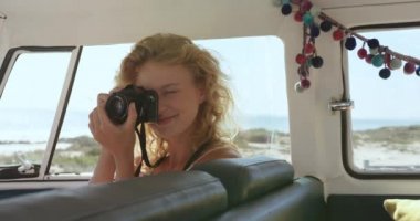 Bir plajda fotoğraf çekmek için bir kamera kullanarak bir kamp minibüsü oturan genç bir Kafkas kadın içinde araç kapatın. Arkadaşları yla plaja Camper Van Yaz Road Trip 4k