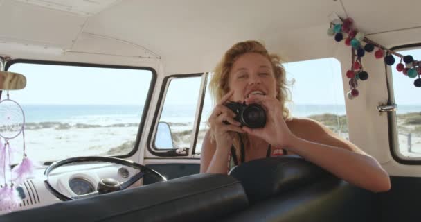 キャンピングカーに乗った若い白人女性が ビーチで写真を撮り カメラに向かって笑いながら キャンピングカーに座っている 友人とビーチにキャンパーバンの夏のロードトリップ4K — ストック動画