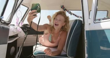 Bir karavanda ön koltuklarda yatan ve kamerayla selfie çeken beyaz bir kadının yakın çekimi. Arkadaşları yla plaja Camper Van Yaz Road Trip 4k