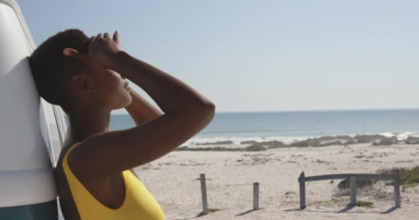 ビーチに駐車したキャンピングカーに身を乗り出した若いアフリカ系アメリカ人女性の横並びをクローズアップし リラックスして太陽を楽しんでいます 友人とビーチにキャンパーバンの夏のロードトリップ4K — ストック動画