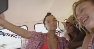 Bir karavanın arkasında oturmuş, akıllı telefonuyla selfie çeken, etnik çeşitliliğe sahip dört genç kız arkadaş. Arkadaşlarıyla birlikte Camper Van 'la sahile doğru yaz gezisi.