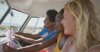 Bir karavanın ön koltuğunda oturan, pencereden dışarı bakıp gülen üç yetişkin arkadaştan oluşan çok ırklı bir grubun yan görüntüsü. Arkadaşlarıyla birlikte Camper Van 'la sahile doğru yaz gezisi.