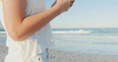 Bir akıllı telefon 4k kullanarak bir plajda duran, güneş şapkası giyen gülümseyen genç Beyaz bir kadının yan görünümüne yakın