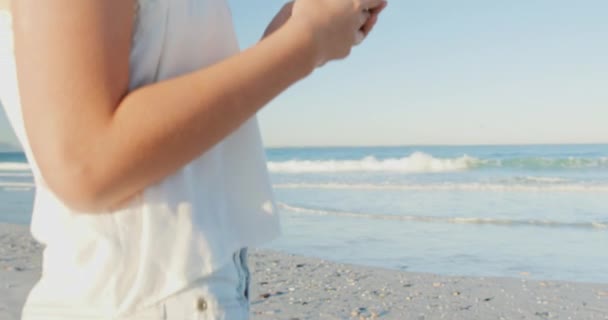 特写一个微笑的年轻白种妇女戴着太阳帽 站在海滩上使用智能手机4K的特写侧视图 — 图库视频影像