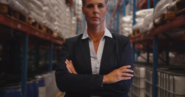 Portrait rapproché d'une femme blanche d'âge moyen gérante d'entrepôt regardant directement à la caméra avec les bras croisés debout par des étagères dans un entrepôt. Un ouvrier d'entrepôt passe devant elle .