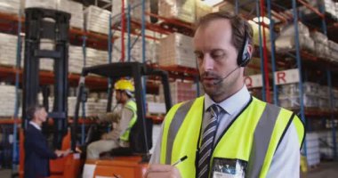 Orta yaşlı beyaz bir erkek depo müdürünü depo yükleme bölümünde iki yönlü telsiz kulaklığı kullanarak kapatın. Arka plandaki depo çalışanları bir forklift kamyonunun yanında konuşuyorlar.. 