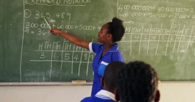 Sınıfın önünde duran, tahtayı işaret eden ve ilkokul sınıfındaki bir ders sırasında sınıfla konuşan Afrikalı bir kızın yan görüntüsü. Ön planda sınıf arkadaşlarının izlediği arka plan.