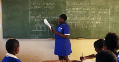 Side view of a young African schoolgirl standing at the front of the class reading from a book she is holding, pointing to the blackboard and talking to the class during a lesson in a township elementary school classroom.
