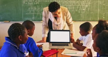 Orta yaşlı bir Afrikalı okul öğretmeninin kasaba ilkokulundaki bir derste öğrencilerine dizüstü bilgisayar göstermesi. Ön planda, masalarda oturan öğrencilerinin dikiz görüntüsü...