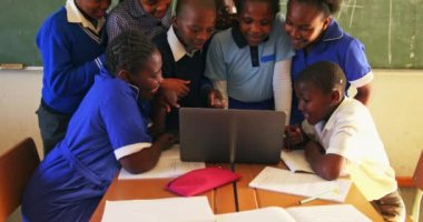 Bir grup Afrikalı okul öğrencisinin, ilkokul 4k sınıfındaki bir ders sırasında bir dizüstü bilgisayarın etrafında toplanıp tahtaya oturdukları görülüyor.
