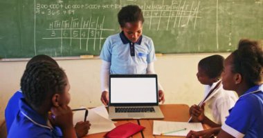 Genç Afrikalı bir öğrencinin, ilkokuldaki bir derste sınıf arkadaşlarına dizüstü bilgisayar gösterirken ön görüntüsü. Ön planda sınıf arkadaşları sıralarında oturup 4K dinliyorlar.