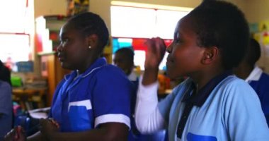 Bir kenar mahalle ilköğretim sınıfındaki bir derse cevap vermek için elini kaldıran Afrikalı genç kızların yan görüntüsü. Arka planda, sınıf arkadaşları dikkatle 4K 'yı dinliyor.