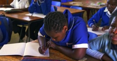 Bir ilkokul dersinde not defterine yazı yazan Afrikalı genç bir kızın ön görüntüsü, arka planda sınıf arkadaşları 4k sorusuna cevap vermek için ellerini kaldırıyorlar.