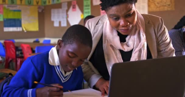 町の小学校の教室でのレッスン中にノートパソコンを使って机の上に座っている若いアフリカの学生を助ける中年のアフリカの女性の学校の先生のフロントビュー — ストック動画
