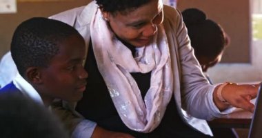Orta yaşlı Afrikalı bir bayan okul öğretmeninin, ilkokul sınıfında ders sırasında dizüstü bilgisayar kullanarak masasında oturan Afrikalı bir öğrenciye yardım edişinin ön görüntüsü.