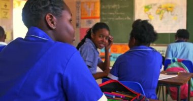 İlkokul 4k sınıfındaki bir derste masalarında oturmuş birbirlerine sınıf arkadaşları hakkında fısıldayan iki Afrikalı genç kızın arka görüntüsü.