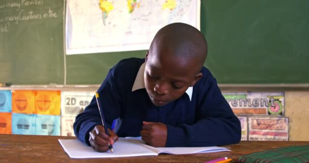 ノートを書きながら机を見上げ町の小学校の教室でのレッスン中に耳を傾けながら机の上に座っている若いアフリカの少年のフロントビュー4K — ストック動画