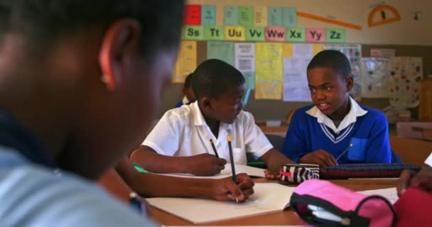 町の小学校の教室での授業中に机に座って話している2人の若いアフリカの学生の正面図 周りのクラスメートも机に座っている4Kを書いている — ストック動画