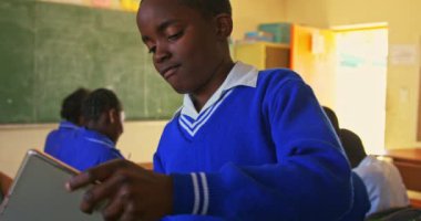 Ön planda masasında oturan Afrikalı bir çocuğun tablet bilgisayara bakıp bir ilkokul sınıfında ders sırasında gülümsemesi var. Arka plandaki sınıf arkadaşları masalarında 4K ile çalışıyorlar.