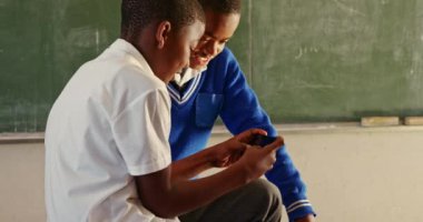 İlkokul 4k sınıfındaki derslere ara verilirken akıllı telefona bakan iki Afrikalı çocuğun yan görüntüsü.